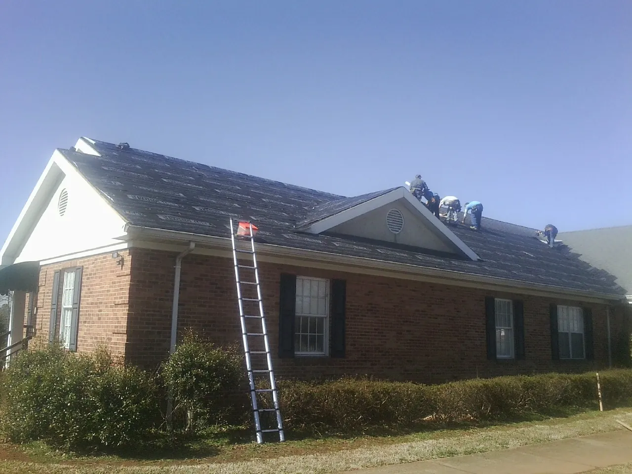 Crew tearing shingles off a brick ranch roof with a ladder set — Emergency Roof Repair work by our crew in Chenango Forks