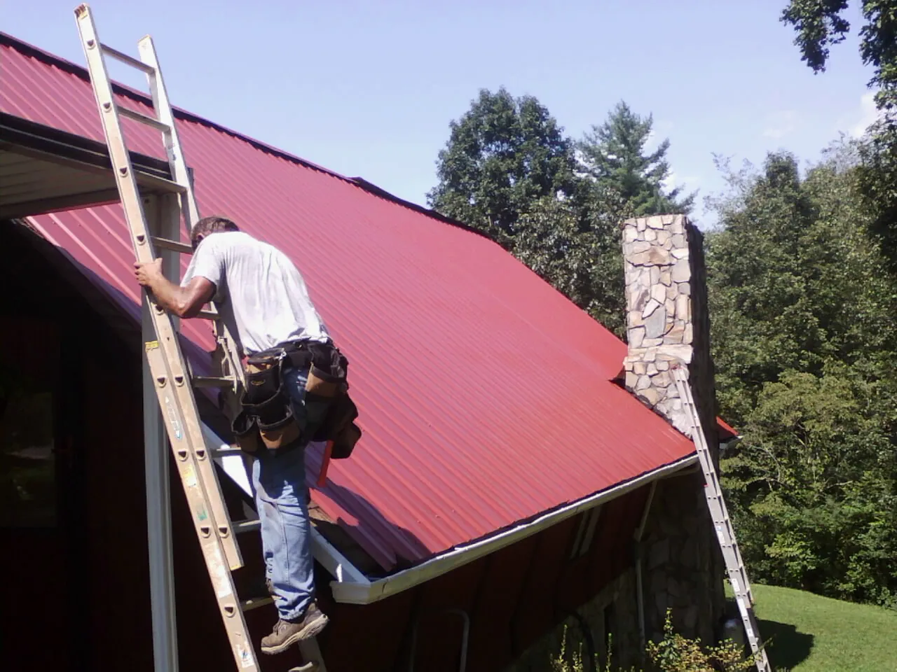 Roofer on a ladder beside a red metal roof and stone chimney — Soffit & Fascia Repair by Roofing Brothers in Chenango Forks